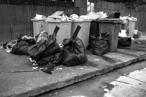 A row of four large wheelie bins positioned on a concrete pavement next to a wooden fence, with the first three bins in light grey and the last in dark grey, all filled with mixed rubbish. Some of the rubbish, including crumpled black plastic bags and loose waste, is piled on the ground in front of the bins, spilling onto the pavement. The waste appears to consist of paper, plastic, and general household or office refuse. The scene suggests an outdoor rubbish collection area, possibly in Archway, with the bins placed on a slightly weathered pavement surface. The surrounding environment includes a wooden fence extending across the background, providing a boundary for the area, and natural lighting indicating daytime conditions. The overall setting aligns with common rubbish removal services offered by companies like Office Clearance Archway for local waste management needs in north London.