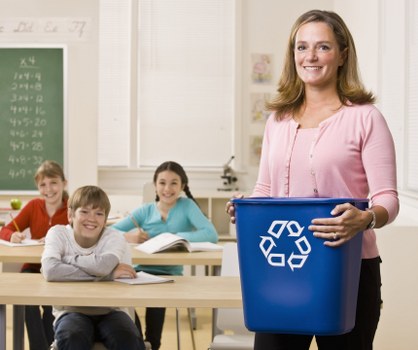 A woman standing in a classroom holding a large blue rubbish bin with a recycling symbol printed on it. Behind her, three children are seated at desks, smiling and facing forward, with one girl wearing a light blue top and two boys wearing white and red tops. The classroom has white walls, a green chalkboard with handwritten notes and figures on the left side, and a window in the background, allowing natural light to illuminate the scene. The woman appears to be part of a waste management or recycling educational activity, with the setting suggesting an environment focused on environmental awareness. The scene reflects a professional approach to waste disposal and recycling, fitting for an office or educational context related to rubbish removal services, such as those offered by Office Clearance Archway in London's N19 postcode area.