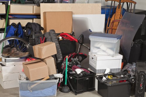 A cluttered indoor scene with a variety of discarded items including cardboard boxes, plastic storage containers, a black toolbox, and miscellaneous household objects, all arranged haphazardly on a concrete or tiled floor. Some boxes are stacked, while others are placed alongside bags, tools, and cables, indicating a recent clearance or decluttering activity. Behind the pile, there are shelves with more stored items, and a glimpse of furniture, such as chairs, can be seen on the right side of the image. The lighting appears natural, possibly coming from nearby windows. The scene reflects an environment typical for rubbish removal services by companies like Office Clearance Archway, servicing residential or commercial properties in the London borough, such as the N19 postal district. The assortment of materials includes cardboard, plastic, and metal, emphasizing the need for professional waste collection and recycling efforts in the London area.