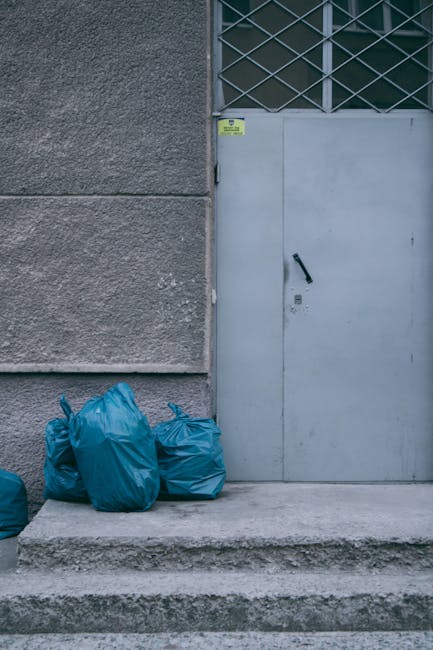 A large, rectangular metal waste skip with a weathered red finish, positioned against a dark green wall and a grey concrete wall in an urban outdoor setting. The skip is partially opened, revealing th
