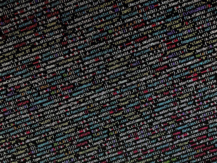 A close-up view of a person's hand typing on a black keyboard positioned on a white desk, with a large computer monitor displaying lines of blue code or text in the background. The workspace includes