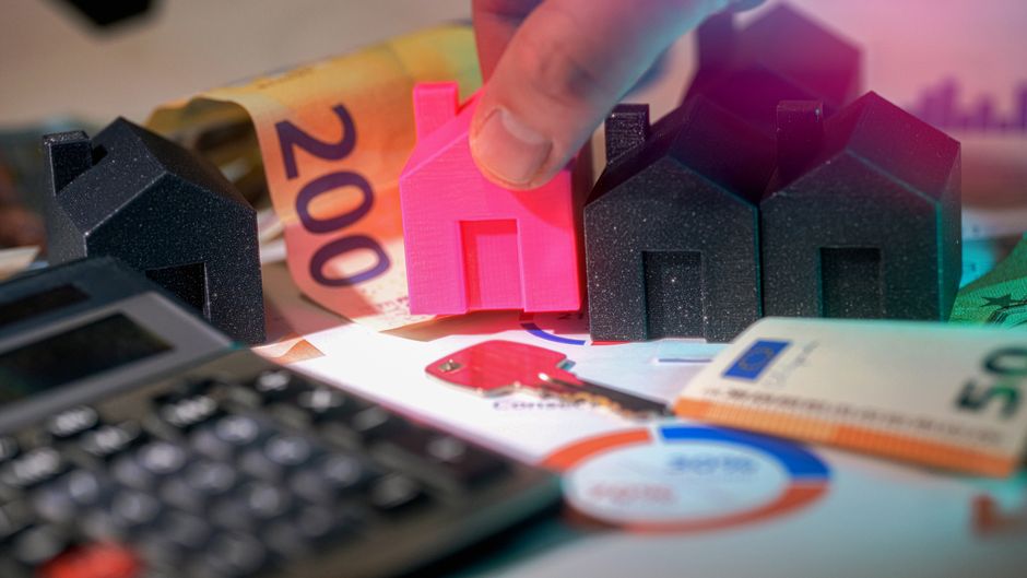 Close-up of a person's hand holding a small, bright pink 3D-printed house model, positioned among three larger black foam houses and a rolled devalued 200 Euro banknote in the background. The scene is